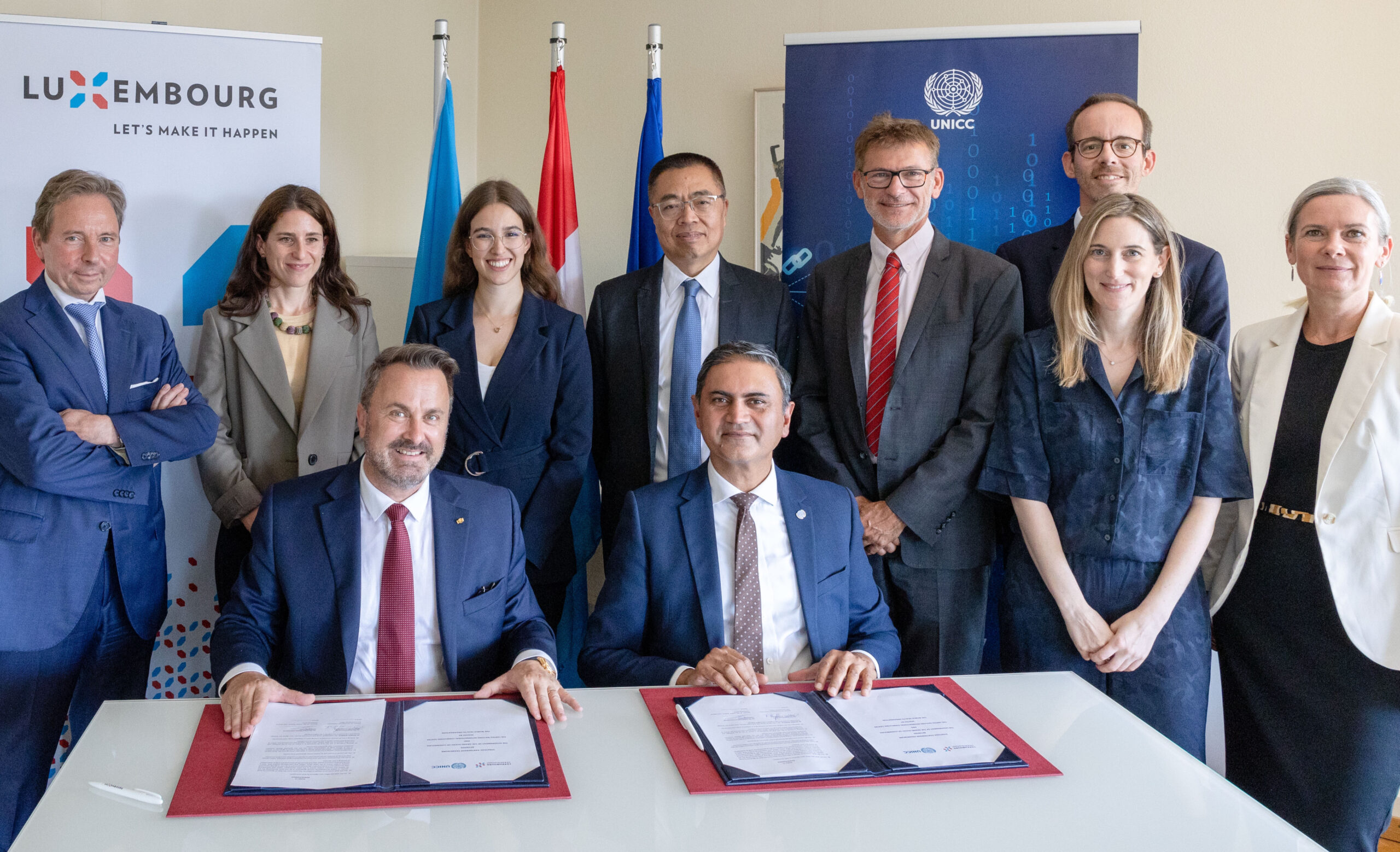 A group of officials and representatives from Luxembourg and UNICC stand and sit together for a photo. Two men seated at a table in the front have signed documents in front of them. Banners behind them feature the Luxembourg logo and the UNICC logo.