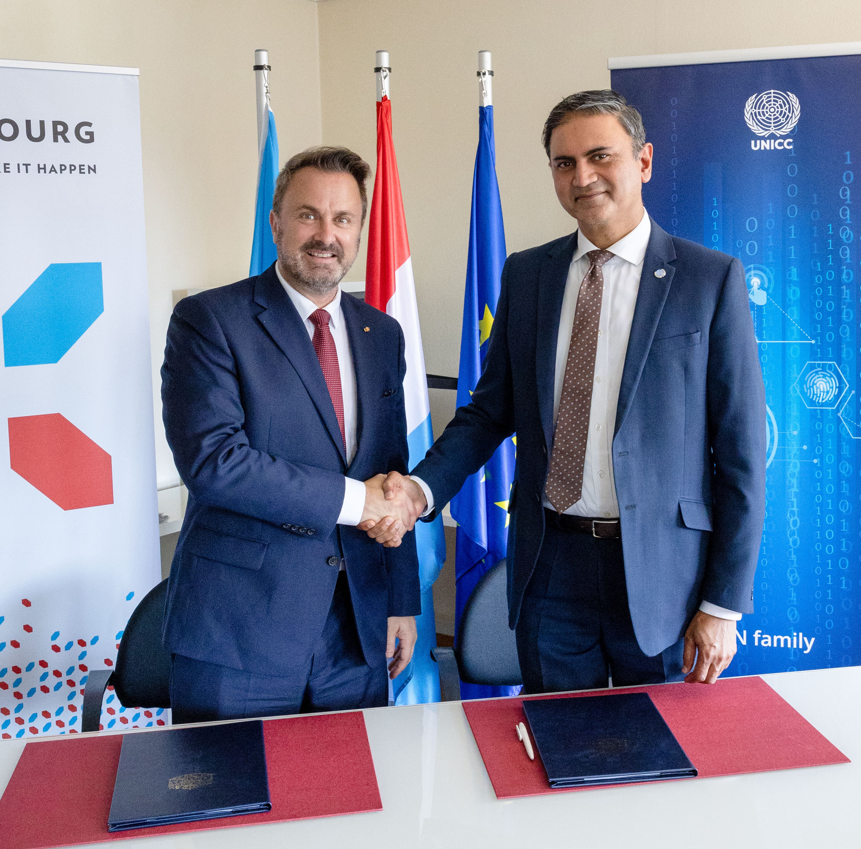 Two men in suits shake hands over a table with documents, representing Luxembourg and the UNICC. Behind them are the flags of Luxembourg and the European Union, and banners for Luxembourg and the United Nations International Computing Centre (UNICC).