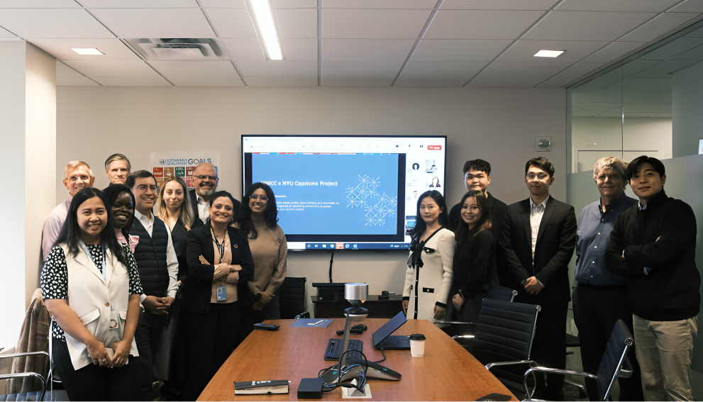 A diverse group of professionals and students posing in a conference room for the UNICC x NYU Capstone Project presentation. A screen in the background displays the project title with a blue slide, and a Sustainable Development Goals poster is visible on the wall. Participants are smiling and dressed in business casual attire, with a mix of in-person and remote attendees shown on the screen.